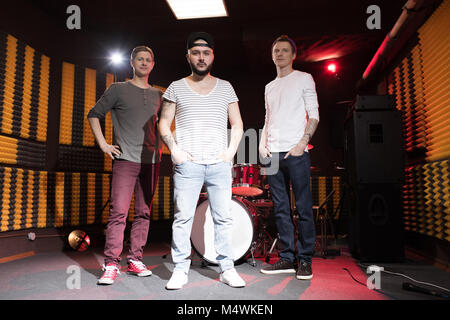 Portrait de groupe de musique moderne posant en studio d'enregistrement et à la recherche à l'appareil photo avec un ensemble de batterie en arrière-plan Banque D'Images