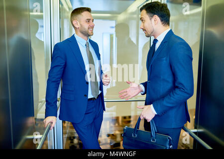 Portrait de deux hommes d'affaires prospères bavardant gaiement équitation ascenseur en verre dans immeuble de bureaux modernes Banque D'Images