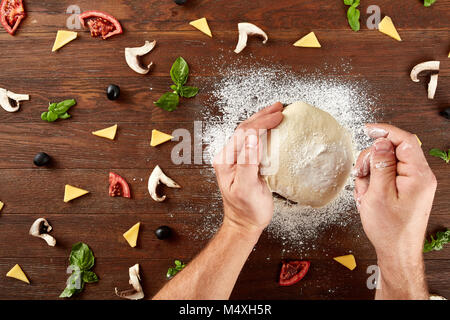 Close-up la main d'un pétrir et façonner la pâte Baker pour la pizza sur une table en bois brun foncé, encadré par quelques morceaux de tomates, champignons, fromage, Banque D'Images