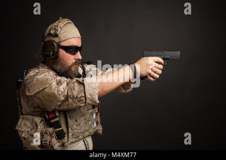 L'homme brutal dans le désert et l'uniforme militaire l'armure de corps debout dans un rack de combat et visant de son arme sur un fond noir. Banque D'Images