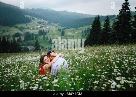 Portrait romantiques en plein air. Pretty woman est heureusement hugging son petit ami tout en étant assis sur le chamonile prairie au fond des montagnes vertes. Banque D'Images