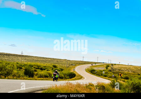 Un motocycliste rides sur une route sinueuse que les courbes de l'avant et vers le haut, rien que des prairies sur chaque côté et un grand ciel bleu au-dessus. Banque D'Images