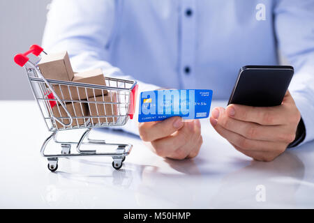 Main tenant une carte de crédit et un téléphone cellulaire pour faire des achats en ligne avec Miniature Shopping Trolley Banque D'Images
