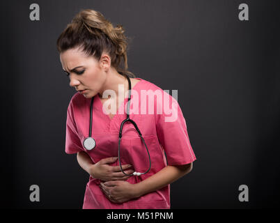Portrait de belle jeune doctor holding estomac comme mal sur fond noir avec copyspace zone publicitaire Banque D'Images