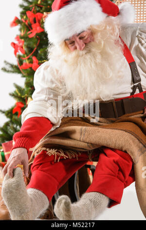 Père Noël assis dans un rocking-chair près de l'arbre de Noël à la maison et des massages son pied Banque D'Images