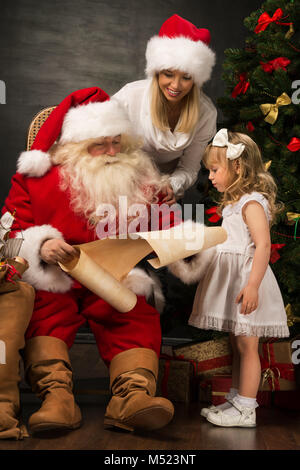 Père Noël assis à la maison avec jolie petite fille et sa mère et la lecture de lettre Banque D'Images