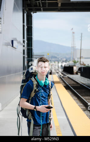 Adolescent, avec un sac à dos sur son dos, écouteurs autour de son cou et la tenue d'un smartphone, attend le train à la gare d'Anaheim. Banque D'Images