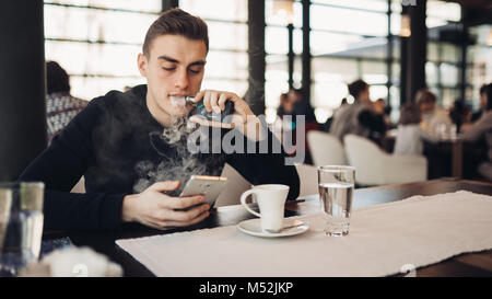 Jeune homme à l'aide de la cigarette électronique pour fumer dans un espace public fermé.satisfait e- cigarette utilisateur dans cafe.interdiction de fumer, la dépendance à la nicotine de substitution,qu Banque D'Images