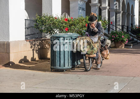 Un homme sans-abri sur son vélo chargé avec ses possessions en scrounging est trash et bacs de recyclage devant l'Hôtel de Ville à Santa Barbara, Cal Banque D'Images