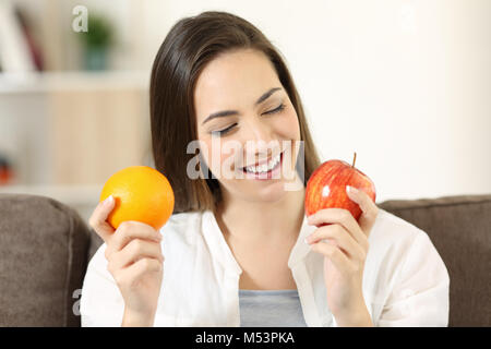 Vue avant portrait d'une femme heureuse de décider entre orange et apple assis sur un canapé dans la salle de séjour à la maison Banque D'Images