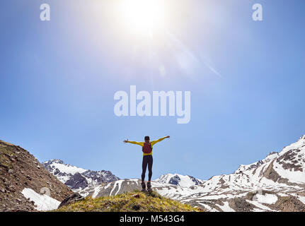 Randonneur dans une chemise jaune avec sac à dos, debout sur le rocher avec l'augmentation des mains en profitant de la vue des montagnes enneigées à sunny day sky Banque D'Images