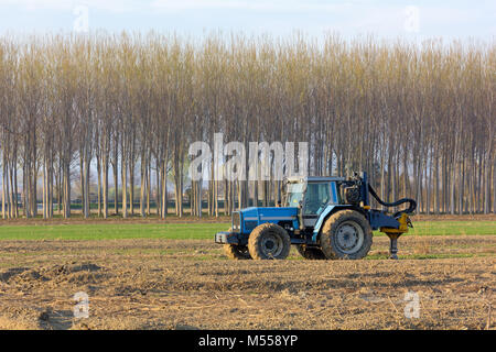 Tracteur bleu au milieu d'un champ avec une rangée d'arbres et une route à l'arrière-plan Banque D'Images