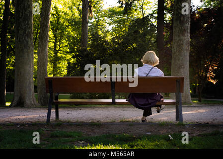 Vieille femme lisant sur un banc dans un parc au coucher du soleil. Elle semble calme et apprécie le soleil. Banque D'Images