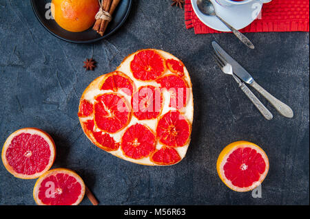 Vue de dessus du gâteau au fromage cuit au four avec des tranches de pamplemousse rouge en forme de cœur sur le fond de la pierre noire avec des ingrédients et des ustensiles. Focus sélectif, de l'espace pour le texte. Banque D'Images