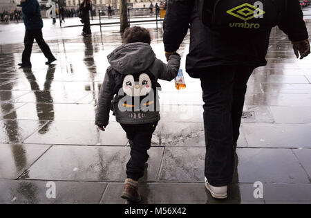 Un jeune garçon tient la main avec un gardien portant un sac à dos pingouin marche à travers les rues humides de Londres. Banque D'Images