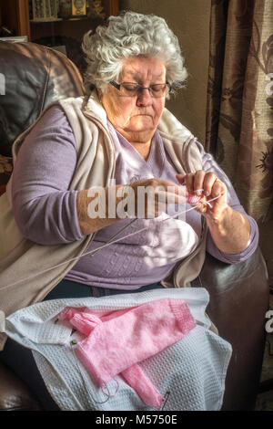 Aux cheveux gris, 80 ans, femme, assise dans un fauteuil en cuir par fenêtre à la lumière du soleil pommelé, un tricot de laine rose / vêtement de laine. Angleterre, Royaume-Uni. Banque D'Images