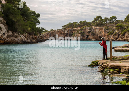 Une jeune femme brune dans un chandail rouge, foulard et cheveux grisonnants prend des photos avec son téléphone portable depuis les tribunes de bateaux de pêche dans la région de Cala Pi de th Banque D'Images