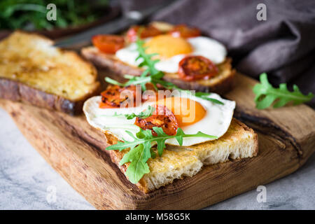 Petit-déjeuner sain avec des œufs, du pain grillé et tomates rôties roquette sur planche à découper en bois rustique. Vue rapprochée Banque D'Images