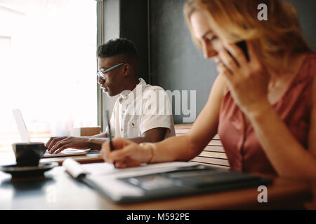 Femme entrepreneur écrit dans le bloc-notes tout en parlant au téléphone mobile. Man working on laptop sitting dans un café. Banque D'Images