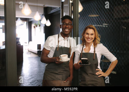 Barista, hommes et femmes se tenant à l'entrée de coffee shop holding tasses à café. Heureux propriétaires de café qui posent avec les tasses de café dans la main. Banque D'Images