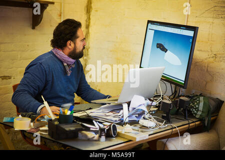 Homme barbu assis à un bureau malpropre dans un atelier, regarder un écran d'ordinateur et un ordinateur portable à l'image d'un couteau. Banque D'Images