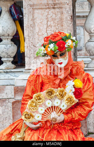 Carnaval de Venise, Venise, Vénétie, Italie. Femme à l'orange vif avec un ventilateur floral or posant avec en détail architectural de la Place St Marc Campani Banque D'Images