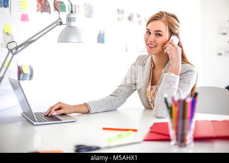 Portrait of young businesswoman using cell phone and laptop in office Banque D'Images