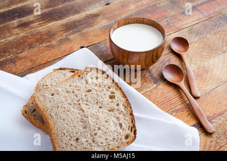 Tranches de pain sur une serviette de cuisine blanc en bois, tasse de lait et deux cuillères en bois sur la diagonale old weathered planches en bois. Banque D'Images