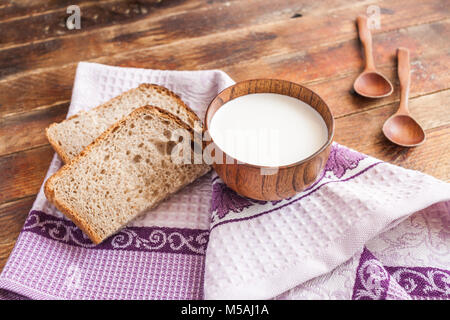 Il y a une tasse de lait, les tranches de pain et deux cuillères en bois sur dishclothon lilas old wooden planks diagonal. Banque D'Images