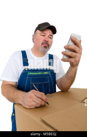 Travailleur en bleu de travail lecture d'un message texte sur son smartphone qu'il note les instructions pour une grande boîte en carton qu'il s'appuie sur isolated on white Banque D'Images
