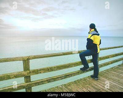 La pensée de l'homme. Séjour touristique en vêtements chauds sur la mole à rampes en bois. Jour brumeux de l'automne. Tourisme à port. Mole, plancher en bois humide au-dessus de la mer. Banque D'Images