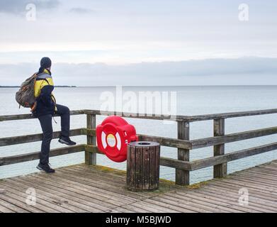 L'homme voyageur avec sac à dos vert assis sur la jetée en bois sur la mer. L'homme en costume de trekking dans la région de harbor en jour brumeux. Mole touristique, plancher en bois humide au-dessus de la mer. Banque D'Images