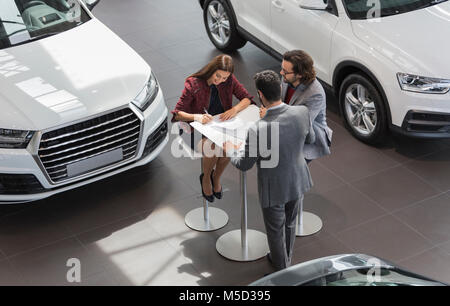 Vendeur de voiture regarder deux clients signature du contrat financier dans la paperasserie car dealership showroom Banque D'Images