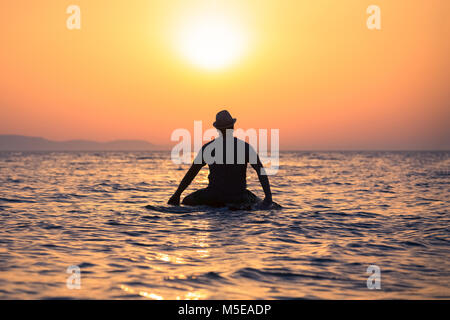 Vue arrière de l'gars avec hat sitting on surfboard dans ocean at sunset Banque D'Images