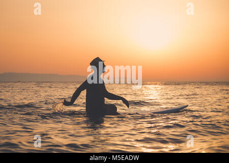 Touriste avec hat sitting on surfboard dans ocean at sunset Banque D'Images