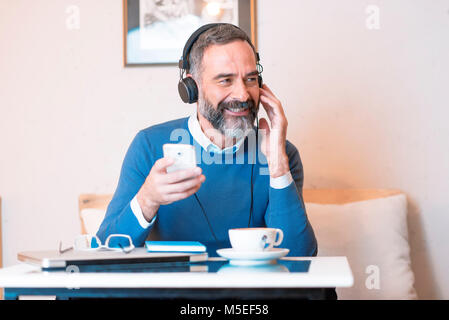 Vieil homme Senior à écouter sa musique préférée au moyen de gros écouteurs, profitant de la richesse du son de la musique, il est assis dans un café Banque D'Images