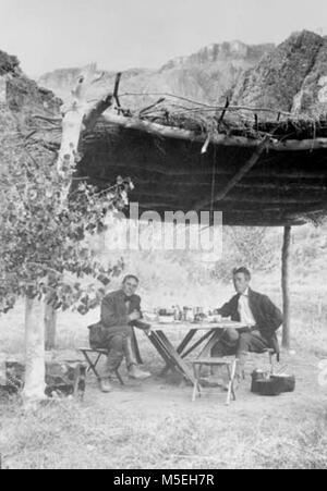 Grand Canyon - Historique Camp rouille c 2 HOMMES ASSIS À TABLE SOUS LE COUVERT DE CHAUME AU CAMP DE SALLE À MANGER LA ROUILLE. Au-delà des falaises. Vers 1910. COLLECTION DE LA ROUILLE. Banque D'Images