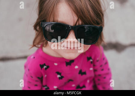 Un bébé fille portant un chandail rose et lunettes, debout sur un trottoir sur une journée chaude et ensoleillée. Banque D'Images