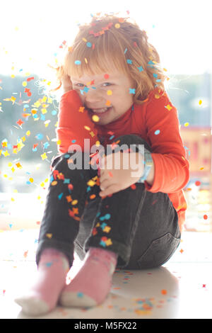 Petite fille timide aux cheveux rouges assis sous l'éclat des confettis sur son anniversaire, souriant et s'amusant. Banque D'Images