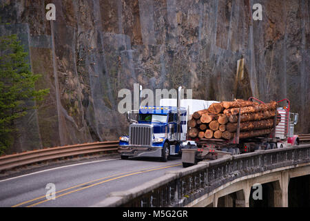 Deux différents modèles de camions semi big rigs classique le transport de billes et une autre cargaison commerciale déménagement l'un vers l'autre rencontré sur le pont au-dessus de t Banque D'Images