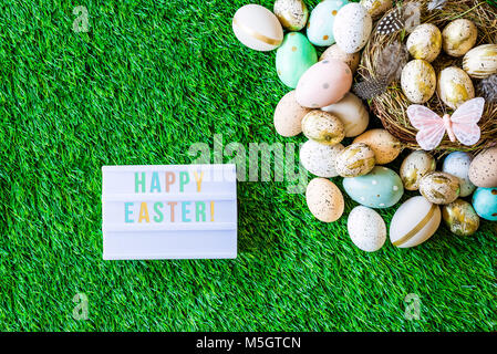 Tourné à partir de ci-dessus comme flatlay avec Happy Easter message écrit sur caisson lumineux et colorés des oeufs de pâques rétro, l'herbe, papillon et nid de Pâques Banque D'Images