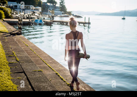 Rear view of woman in sportswear marcher avec un tapis de yoga au Lakeshore Banque D'Images