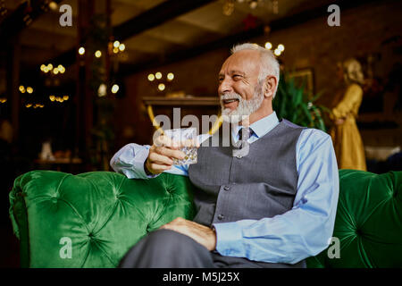 Portrait d'élégante senior man sitting on couch in a bar holding tumbler Banque D'Images