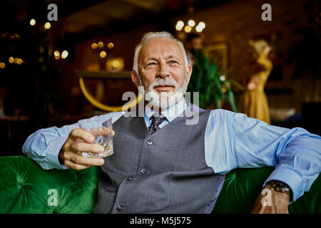 Portrait d'élégante senior man sitting on couch in a bar holding tumbler Banque D'Images