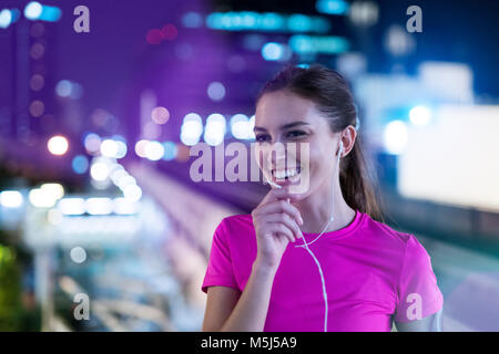 Smiling young woman in pink sportshirt en écoutant de la musique en ville la nuit Banque D'Images