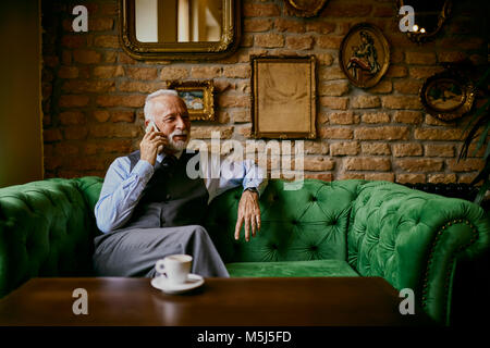 Élégant senior man sitting on couch in a cafe talking on cell phone Banque D'Images