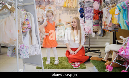 Girl's Shopping - petite fille blonde avec maman choisit robe a la mode dans le magasin de vêtements pour enfants Banque D'Images