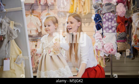 Shopping pour les enfants - petite fille blonde avec robe maman choisit dans le magasin de vêtements pour enfants Banque D'Images