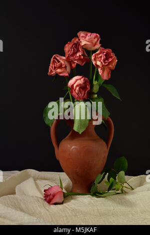 Cinq roses roses mourir dans un vase en terre cuite sur un fond noir, avec une seule rose sur tissu de mousseline. Banque D'Images