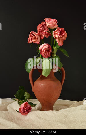 Cinq roses roses mourir dans un vase en terre cuite sur un fond noir, avec une seule rose sur tissu de mousseline. Banque D'Images